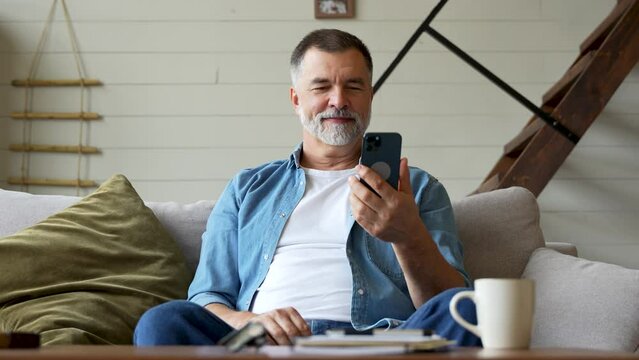 Smiling senior grey-haired man sitting on couch and using mobile phone at home, chatting with his family or using mobile application