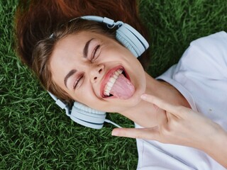 Teenage girl listening to music in headphones lying on the green grass in the park and smiling and showing her tongue in a white T-shirt, summer mood as a lifestyle