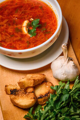 Traditional Borscht Served With Garlic And Bread. Top-down view of a bowl of red beet soup garnished with sour cream and parsley, alongside garlic cloves and toasted bread on a wooden board.