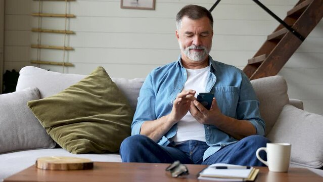 Smiling senior grey-haired man sitting on couch and using mobile phone at home, chatting with his family or using mobile application