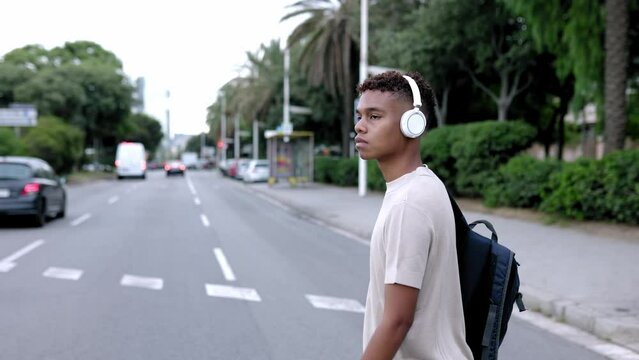 Concept pedestrians passing a crosswalk. Slow motion footage of young confident hispanic latin teenage boy walking across a zebra crossing while listening to music with wireless headphones at city.