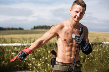 male vandal with a dirty naked torso and a knife in his hand, against the backdrop of an overgrown...