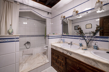 Vintage stately-style bathroom with marble countertops, twin sinks, a large mirror with shades, and a bathtub between white tiles