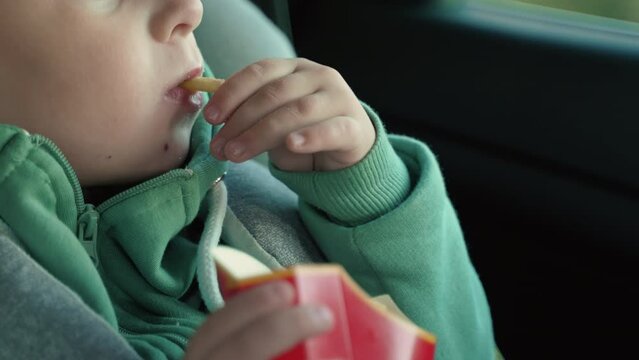 Cropped image of a little boy sitting in a toddler car seat and eating French fries from a packet. High quality 4k footage