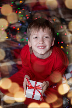 Child boy sitting on a plaid near the christmas tree at home, kid holding in hands a gift box, top view
