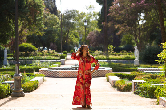 A beautiful woman wears a traditional Moroccan dress in red and embroidered in gold and silver. The girl poses for wedding photos in a Moroccan style garden with fountains and lush greenery.