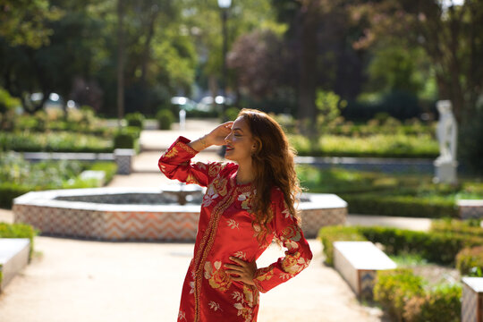 A beautiful woman wears a traditional Moroccan dress in red and embroidered in gold and silver. The girl poses for wedding photos in a Moroccan style garden with fountains and lush greenery.