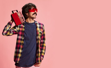 Portrait of stylish man with moustache posing in red sunglasses, checkered shirt and vintage radio player isolated over pink background