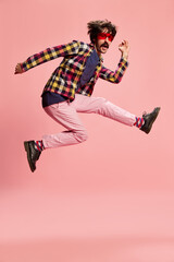 Fototapeta na wymiar Portrait of stylish man with moustache, jumping, posing in checkered shirt and pink pants isolated over pink background