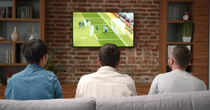 Three men sit on sofa watching football match