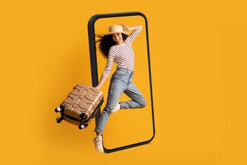 Smiling excited young arab lady tourist in hat with suitcase jumping near huge smartphone, ready...