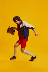 Fototapeta na wymiar Portrait of man with moustache posing in vintage sportswear and red cycling shorts isolated over yellow background. Dancing with vintage music player
