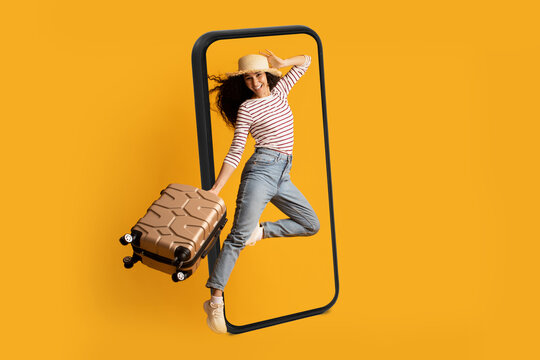 Smiling excited young arab lady tourist in hat with suitcase jumping near huge smartphone, ready for travel