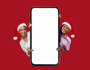 Cheerful black millennial ladies in Santa Claus hats peeking out from big smartphone with empty...