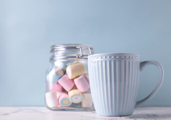 Sweet romantic postcard. Blue cup with hot drink and jar with colorful marshmallows on white marble background against blue wall. Selective focus. Place for text.
