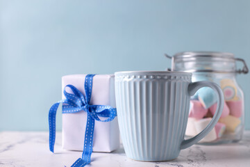 Festive romantic postcard. Blue cup with hot drink, box with present and jar with colorful marshmallows on white marble background against blue wall. Selective focus. Place for text.