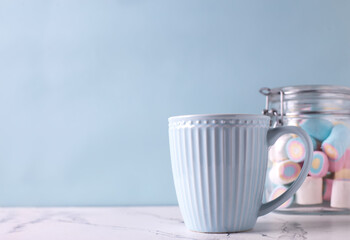 Romantic postcard. Blue mug with hot drink and jar with colorful marshmallows om white marble background against blue wall. Selective focus. Place for text.