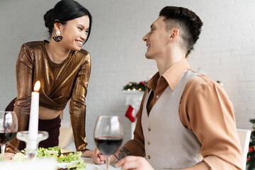 cheerful interracial couple looking at each other near festive meal and glasses of wine during Christmas
