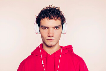 Studio portrait of handsome young gay man with brunette curly hair, wears pink hoodie, listens music with headphones while standing against pastel beige background. Technology concept.