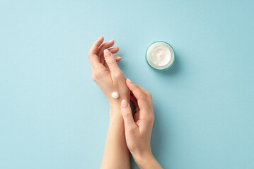 Winter season skin care concept. First person top view photo of young woman applying cream on her hands from cosmetic jar on isolated light blue background