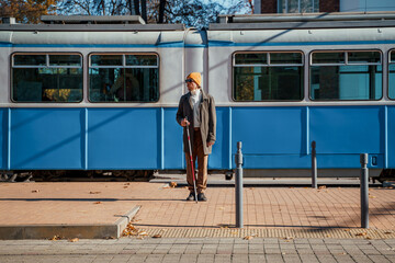 Blind man with a cane stopped on the sidewalk in front of a passing tram