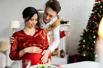 smiling man touching tummy of pregnant asian woman while celebrating Christmas at home