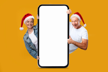 Happy european elderly woman and man in Santa Claus hats show thumb up with big phone with blank...