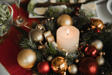 pine wreath with Christmas balls and lit candle on table with festive supper on blurred background
