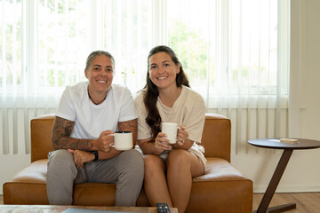 Adult lesbian couple sit on couch with coffee at home