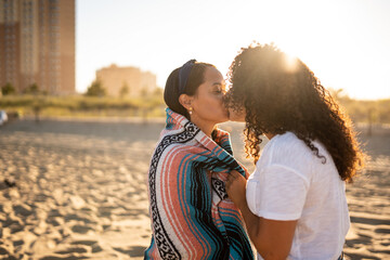 Hispanic lesbian couple kiss while wrapped in blanket at beach