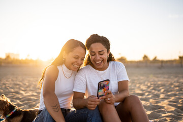 Hispanic lesbian couple use a phone and sit on a blanket at the beach with a dog