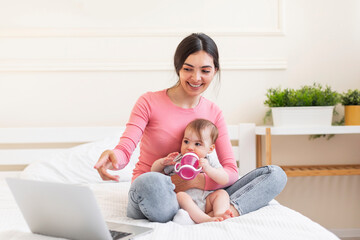 Young mom sitting with baby and making video call via laptop at home, resting on bed and pointing...