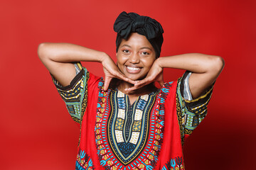 Beautiful black woman with typical dress and turban rests her head on her arms while looking at the camera. She is standing on a red background