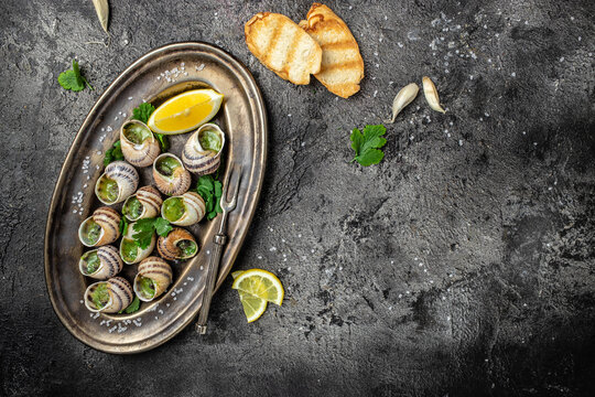 Delicious cooked sea escargo snails with herbs, butter, garlic on metal plate with forks. wine glass. gourmet food. Restaurant menu, Traditional French cuisine