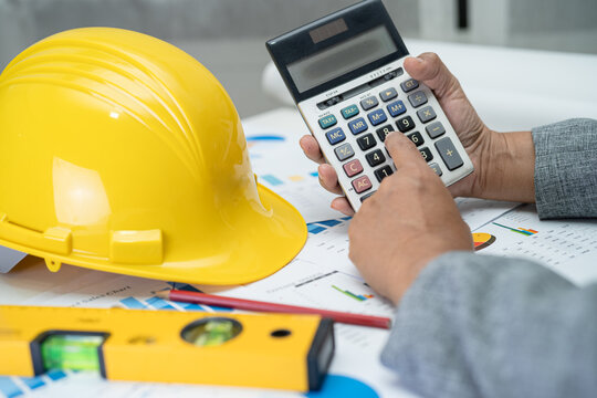 Asian engineer with architectural house plan project blueprint, graph and yellow helmet and engineering construction tools.