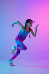 Fototapeta na wymiar Running technique. Sportive girl, junior runner in stylish sportswear and cap posing isolated on gradient pink-blue background with neon filter, light.