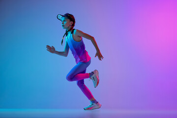 Fototapeta na wymiar Champion. Sportive little girl, junior runner in stylish sportswear and cap running isolated on gradient pink-blue background in neon. Concept of sport, fashion, fitness and education