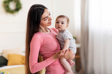 Motherhood concept. Young mom with infant daughter on arms posing at home and smiling, copy space