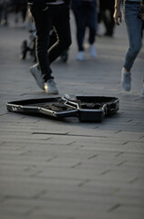 A street musician's black guitar case lies open on the pavement to collect money. Legs and shoes of people can be seen walking around the guitar case.