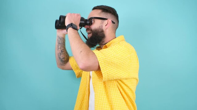 Crazy emotions. Young funny hipster guy with binoculars isolated on blue studio background. Discounts, sales, seasonal sales. Colorful summer concept.