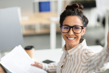 Happy businesswoman having video call or taking selfie while working with documents in office and...
