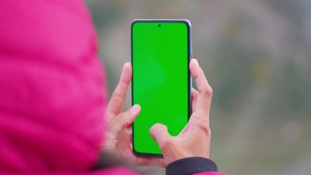 Closeup shot of Indian girl wearing jacket and using mobile phone with Green screen. Green screen mockup template of Mobile phone, Chroma key. Finger swiping up in green screen of the mobile