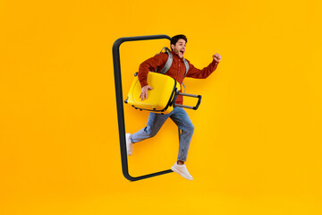 Fototapeta na wymiar Glad excited young european guy tourist with suitcase and backpack jumping out of huge smartphone