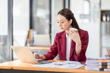 Beautiful young smiling Asian businesswoman working on laptop and drinking coffee, Asia businesswoman working document finance and calculator in her office.
