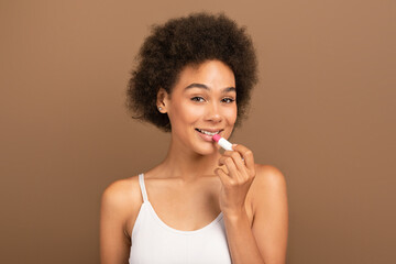 Smiling young african american curly woman in white top apply moisturizing lipstick on lips