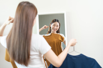 Fototapeta premium Choice of clothes,Nothing to wear. Attractive asian young woman, girl looking into mirror, try on appare, choosing dress, outfit on hanger in wardrobe at home. Deciding blouse what to put on which one