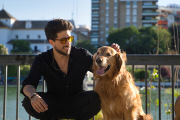 A young Hispanic man with a beard, sunglasses and a black shirt, climbed on a bench with his dog as he caresses him under the sun's rays. Concept animals, dogs, love, pets, golden.