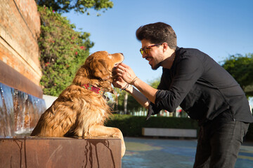 Young Hispanic man with a beard, sunglasses and black shirt, petting his wet dog that has gotten into a fountain because of the heat. Concept animals, dogs, love, pets, golden.