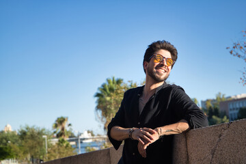 Young Hispanic man with beard, sunglasses and black shirt, looking at the sunset smiling happily, leaning on a stone wall. Model concept, beauty, fashion, triumph, happiness.