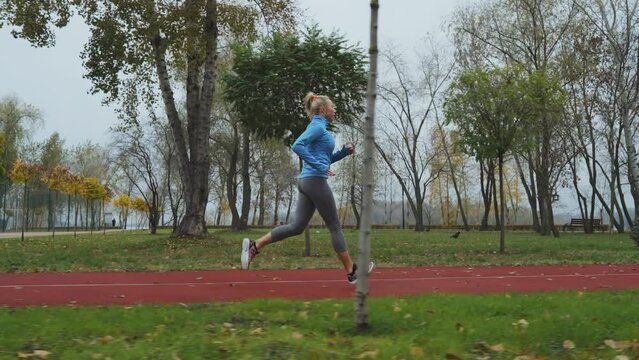 Slow motion fit woman wearing blue sports jacket running on track in autumn park, fallen leaves lying on ground. Tracking shot of active person training in the morning. Concept of fitness